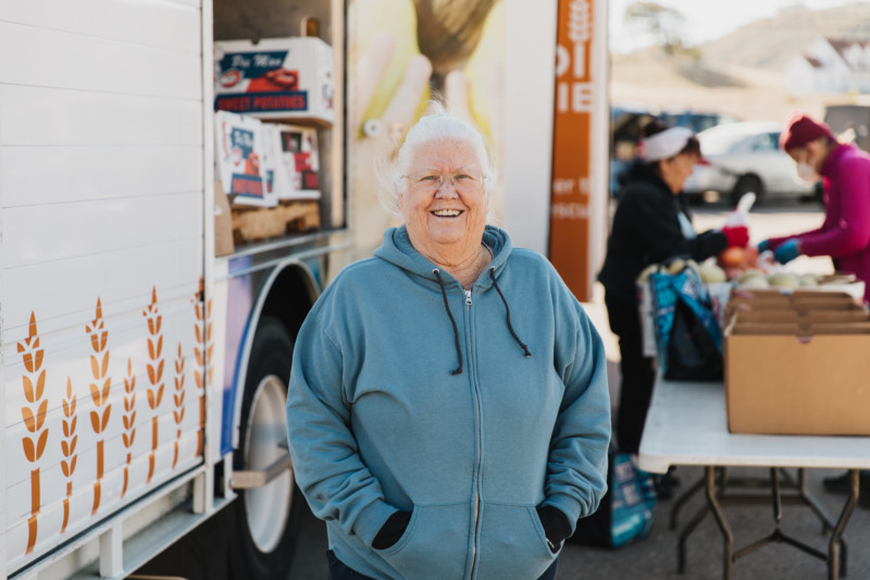A woman with white hair stands in front of a Feeding San Diego truck at a mobile pantry that supports the Feeding Seniors program