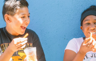 Two kids eat lunch and laugh in front of blue wall