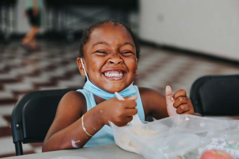 Little girl smiles at camera while eating lunch at a Feeding San Diego summer meals site