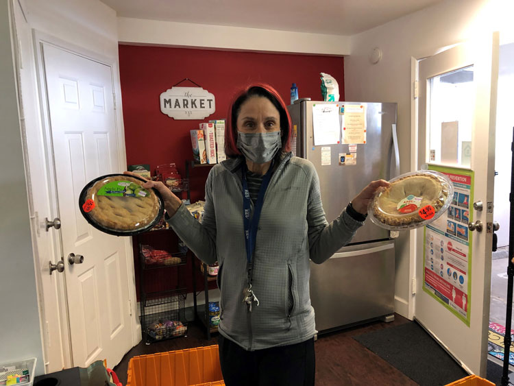 A woman with a face mask and a gray shirt holding pie in each hand in the Wesley House food pantry