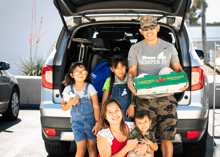 A family of five stands in front of an open truck holding food from a Feeding San Diego distribution