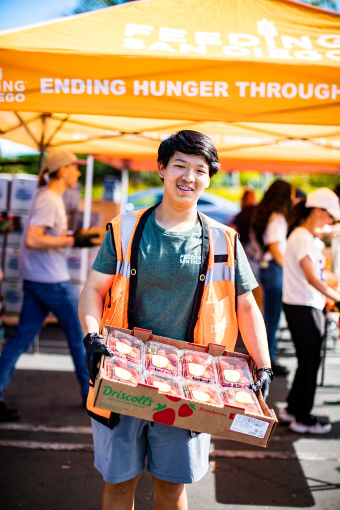 A teenage volunteer with black hair wearing an orange reflective vest and holding a box of strawberries in front of an orange Feeding San Diego canopy