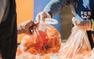 A volunteer wearing a Feeding San Diego blue apron hands a bag with orange peppers to a mobile pantry attendee