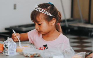 A little girl wearing a tiara eats lunch