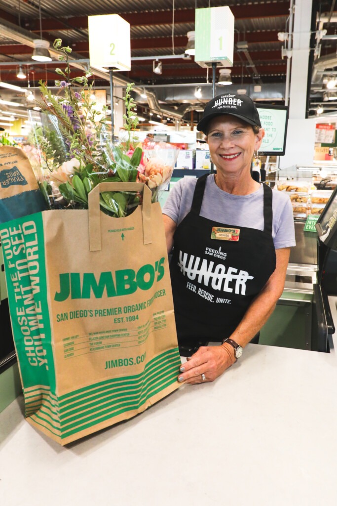 A woman wearing a Cross Out Hunger hat and apron holds a paper bag from Jimbo's