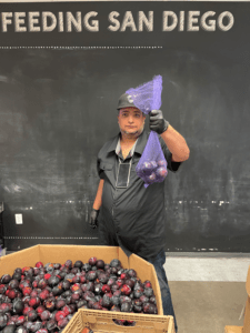 A man in a black baseball cap holds up a bag of plums