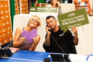 A girl holding a cutout purple heart and a man with a phone up to his ear holding a sign that says "Call me to donate! 858.500.7805"