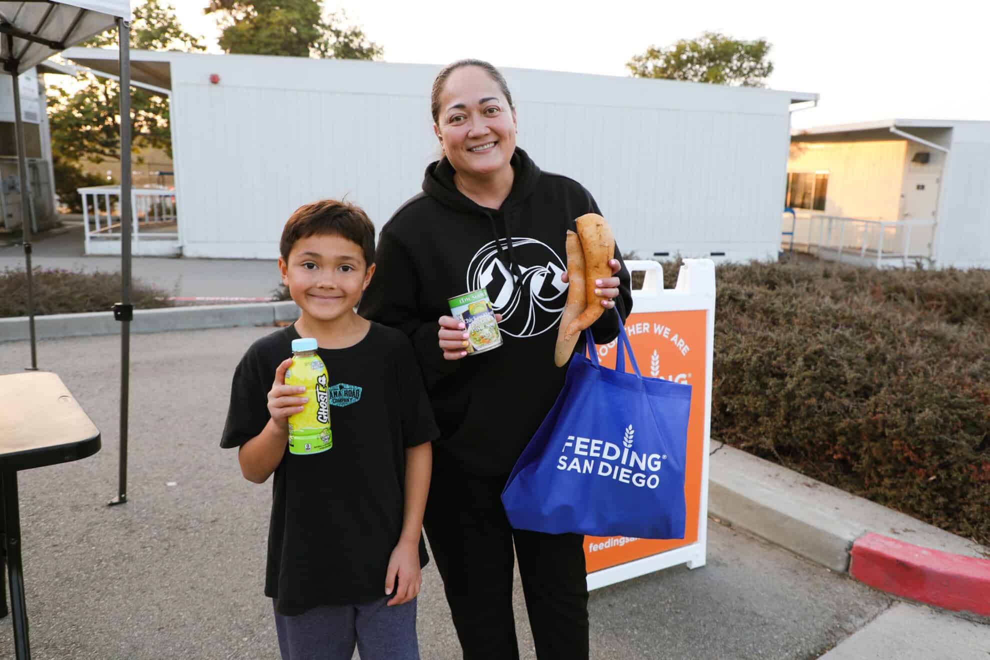 A woman holding produce and a can standing next to a boy holding a bottled drink